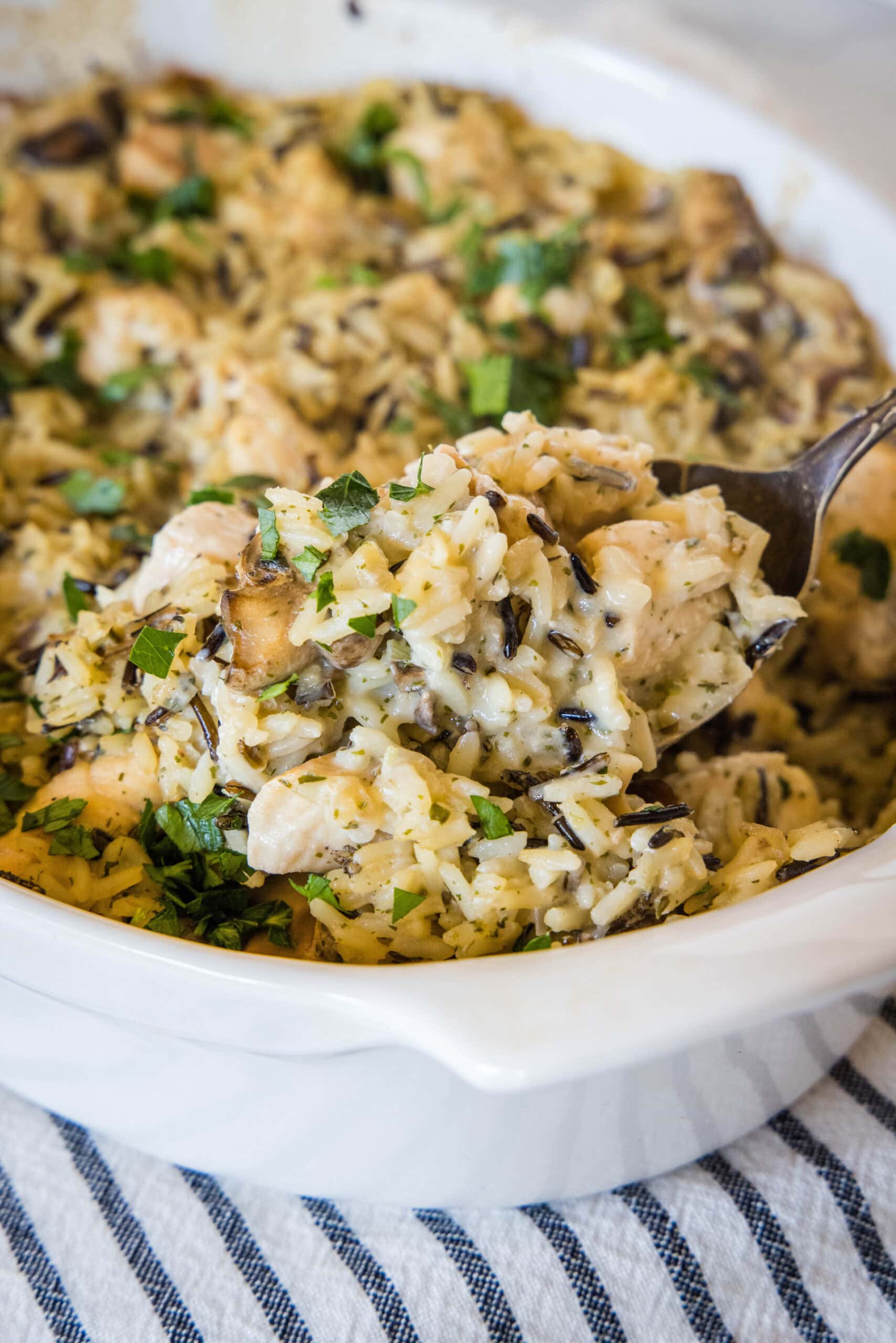 A spoon scooping a serving from chicken wild rice casserole in a ceramic baking dish.