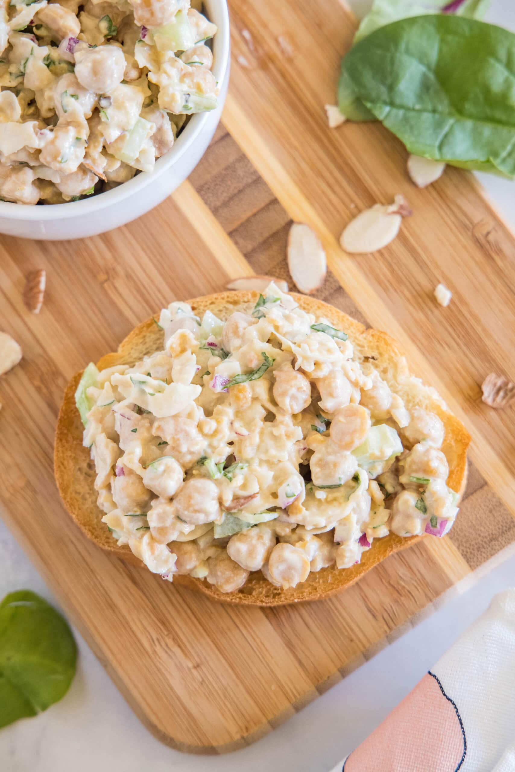 Overhead view of chickpea chicken salad served on a slice of bread next to a bowl of chickpea salad on a wooden cutting board.