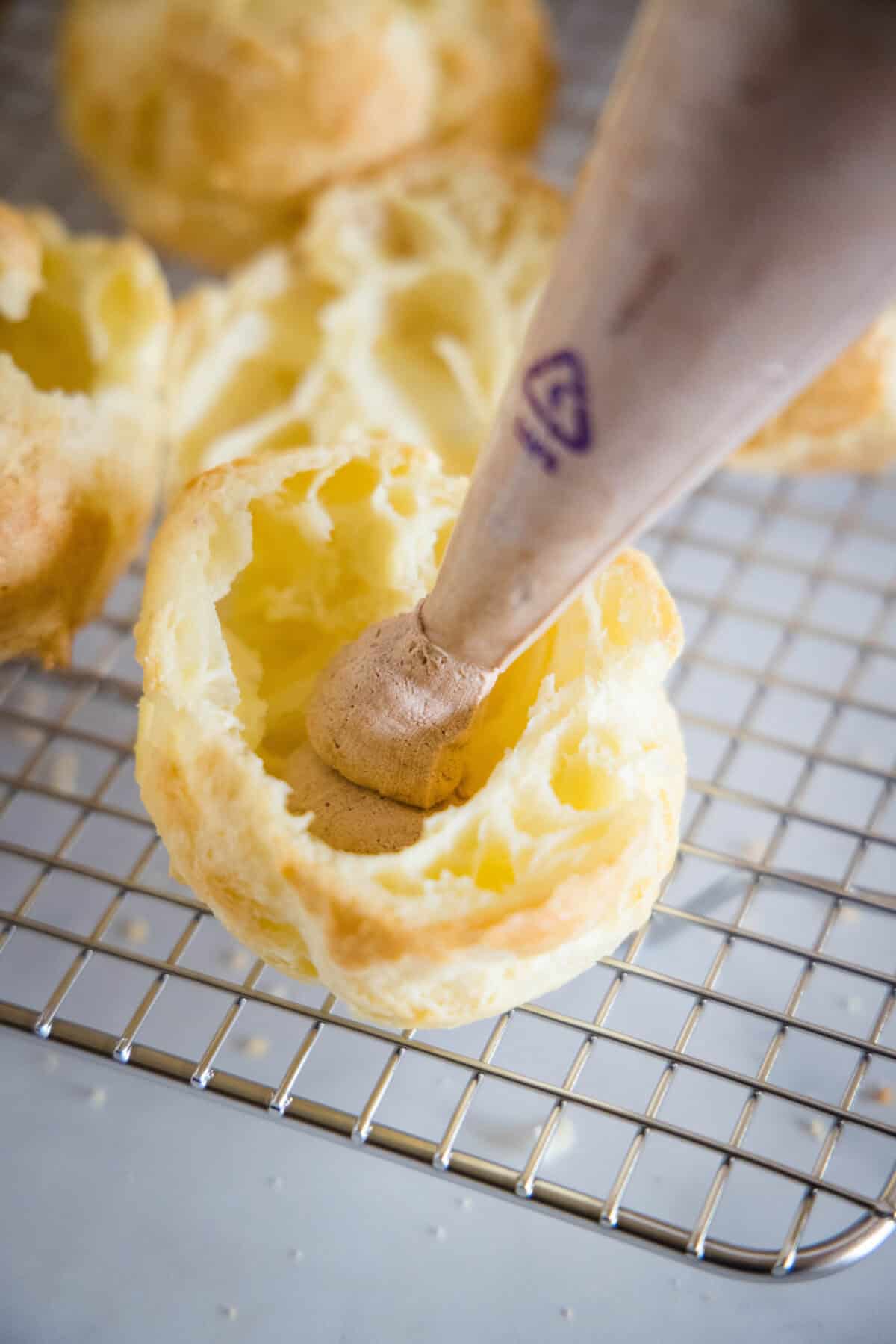 The tip of a piping bag filled with chocolate whipped cream held over an open cream puff on a wire rack.