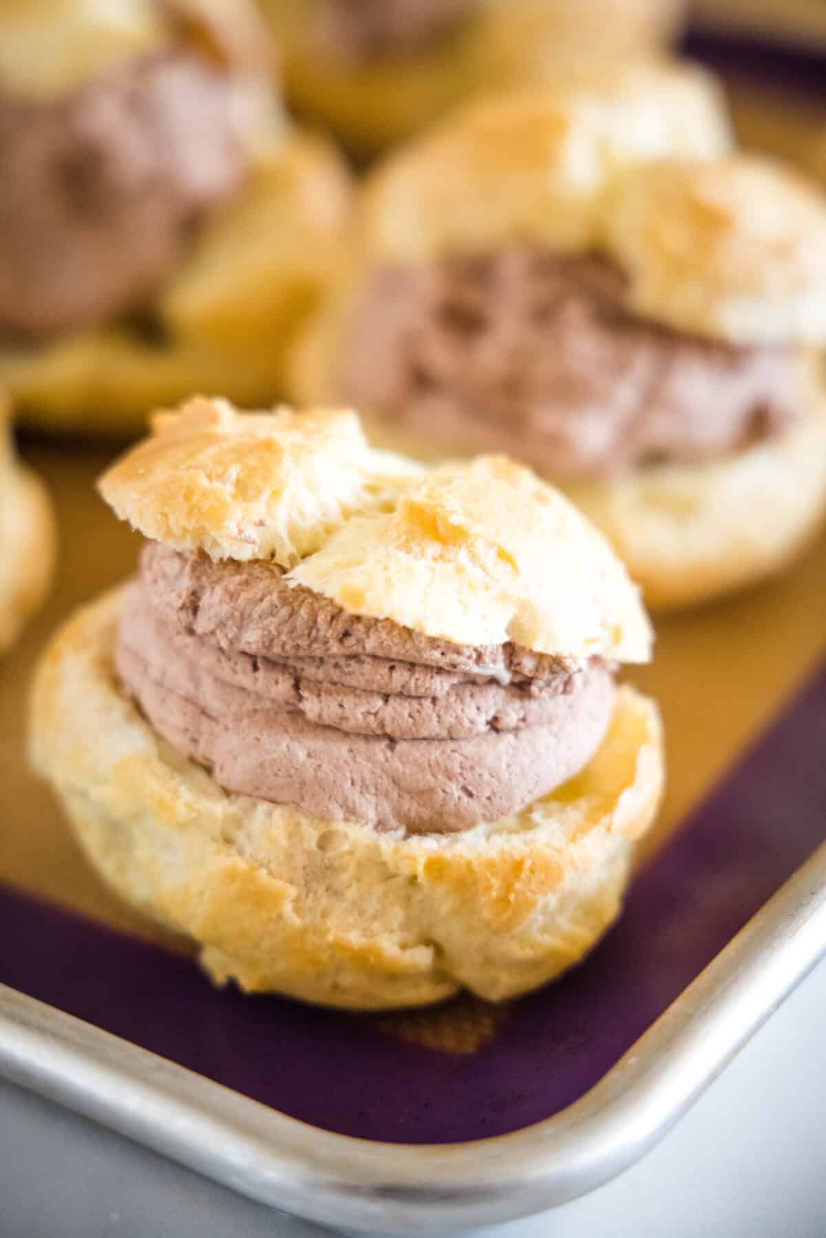 Close up of a filled chocolate cream puff in the corner of a baking pan with more filled puffs in the background.