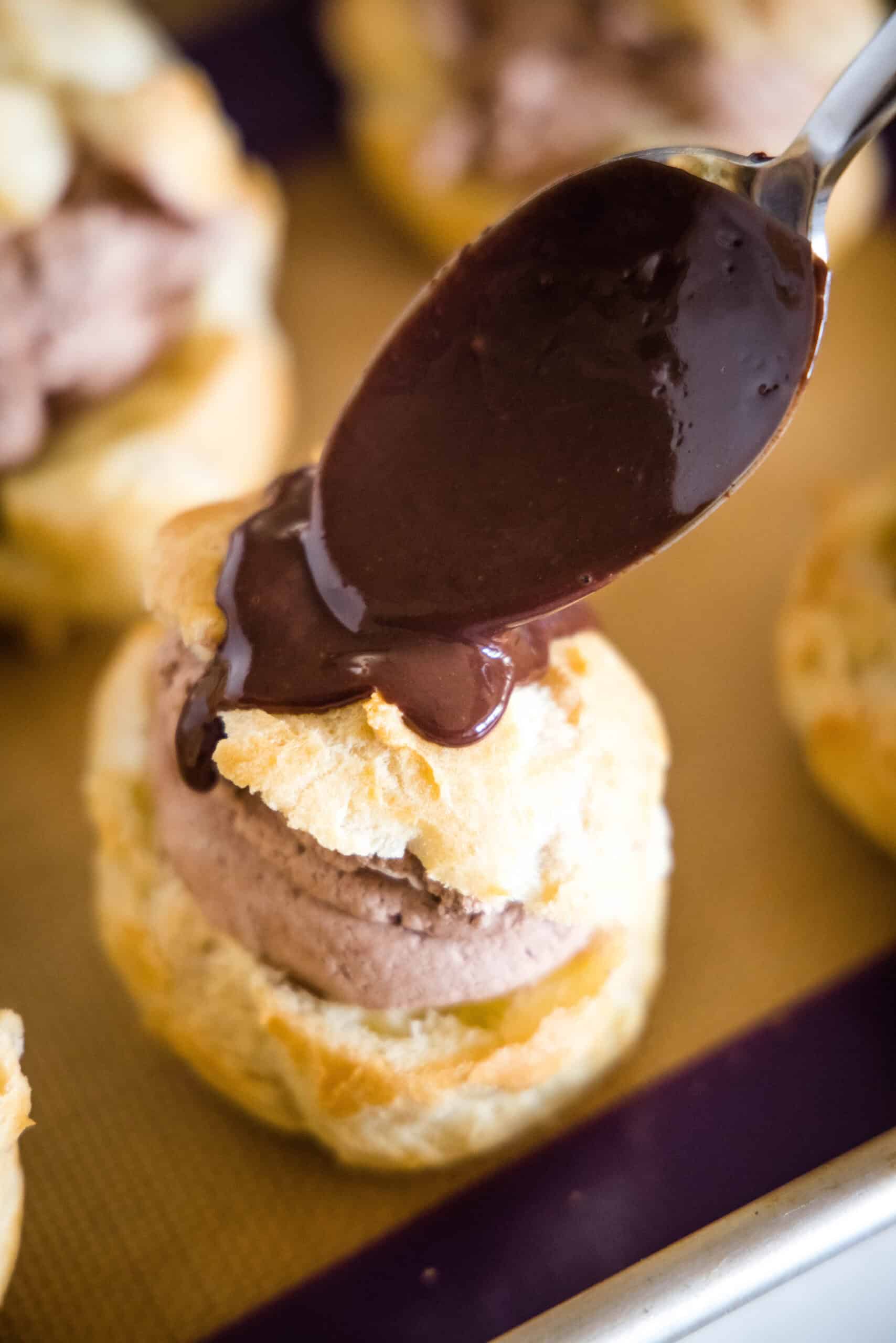 A spoon dripping chocolate ganache on top of a prepared chocolate cream puff.