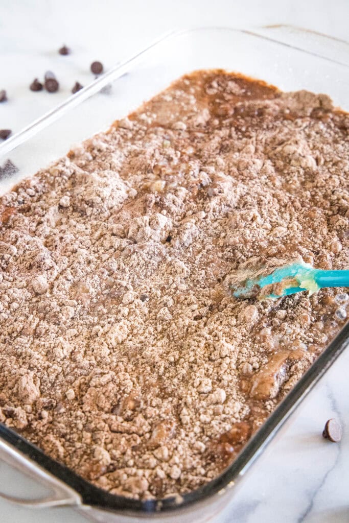 A spatula is used to barely stir the liquid ingredients into dry chocolate cake mix in a 9x13-inch glass baking dish.