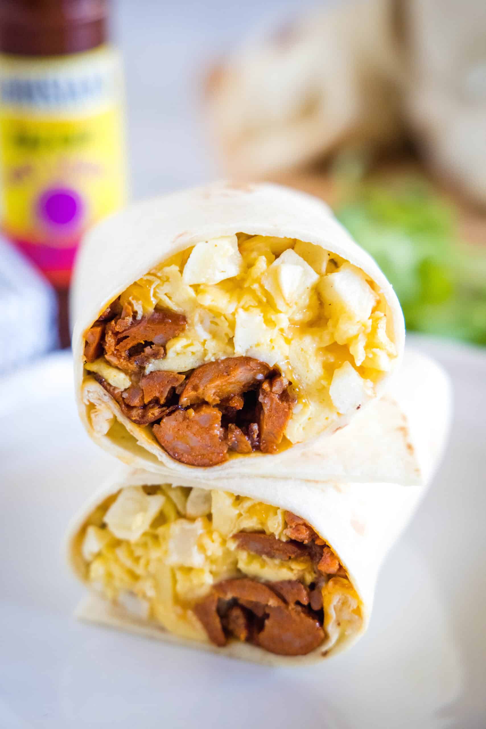 This chorizo breakfast burrito is a cheesy breakfast wrap loaded with crispy hash brown potatoes, spicy chorizo sausage, and fluffy eggs! Two halves of a chorizo breakfast burrito stacked on a plate, showing the eggs, cheese, potatoes, and chorizo filling inside.