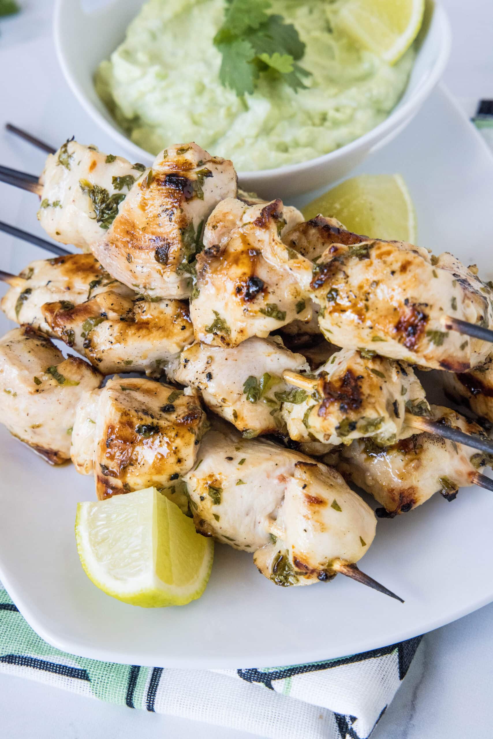 Juicy cilantro lime chicken is an easy recipe filled with bright flavors! Marinated in lime juice with fresh herbs, and grilled on skewers. A plate of grilled cilantro lime chicken skewers next to a lime wedge, with a bowl of avocado dipping sauce in the background.