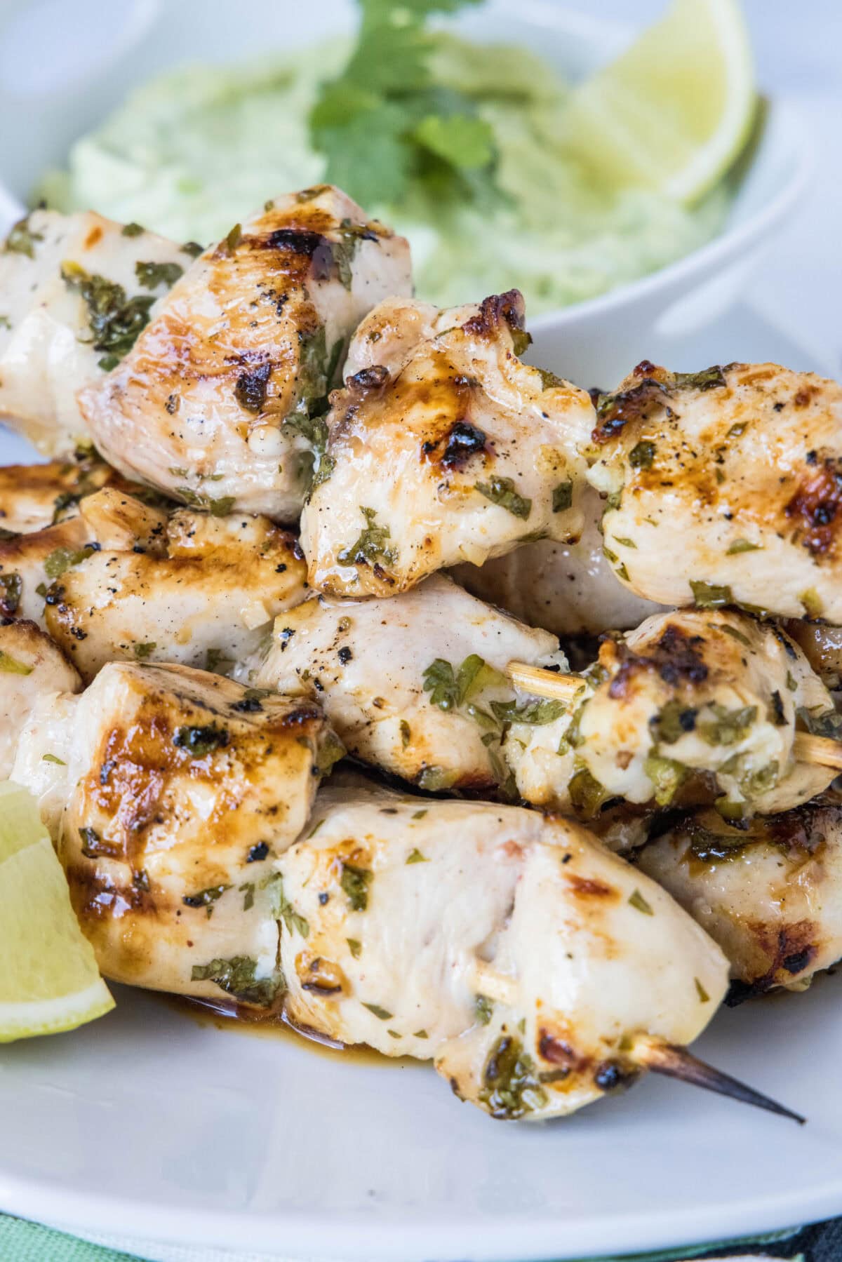 A plate of grilled cilantro lime chicken skewers next to a lime wedge, with a bowl of avocado dipping sauce in the background.