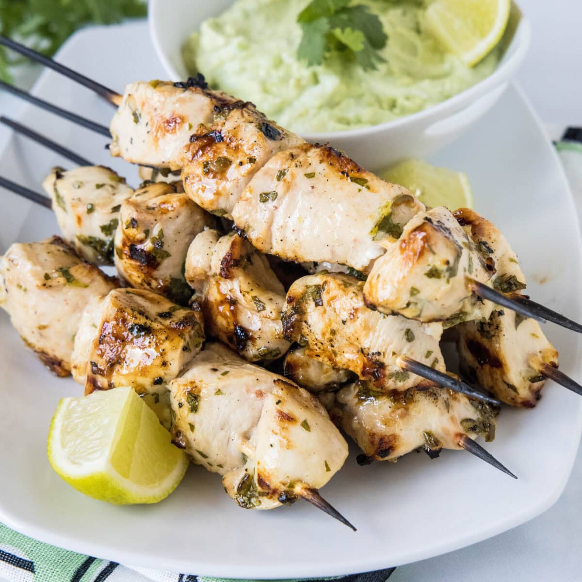 A plate of grilled cilantro lime chicken skewers next to a lime wedge, with a bowl of avocado dipping sauce in the background.