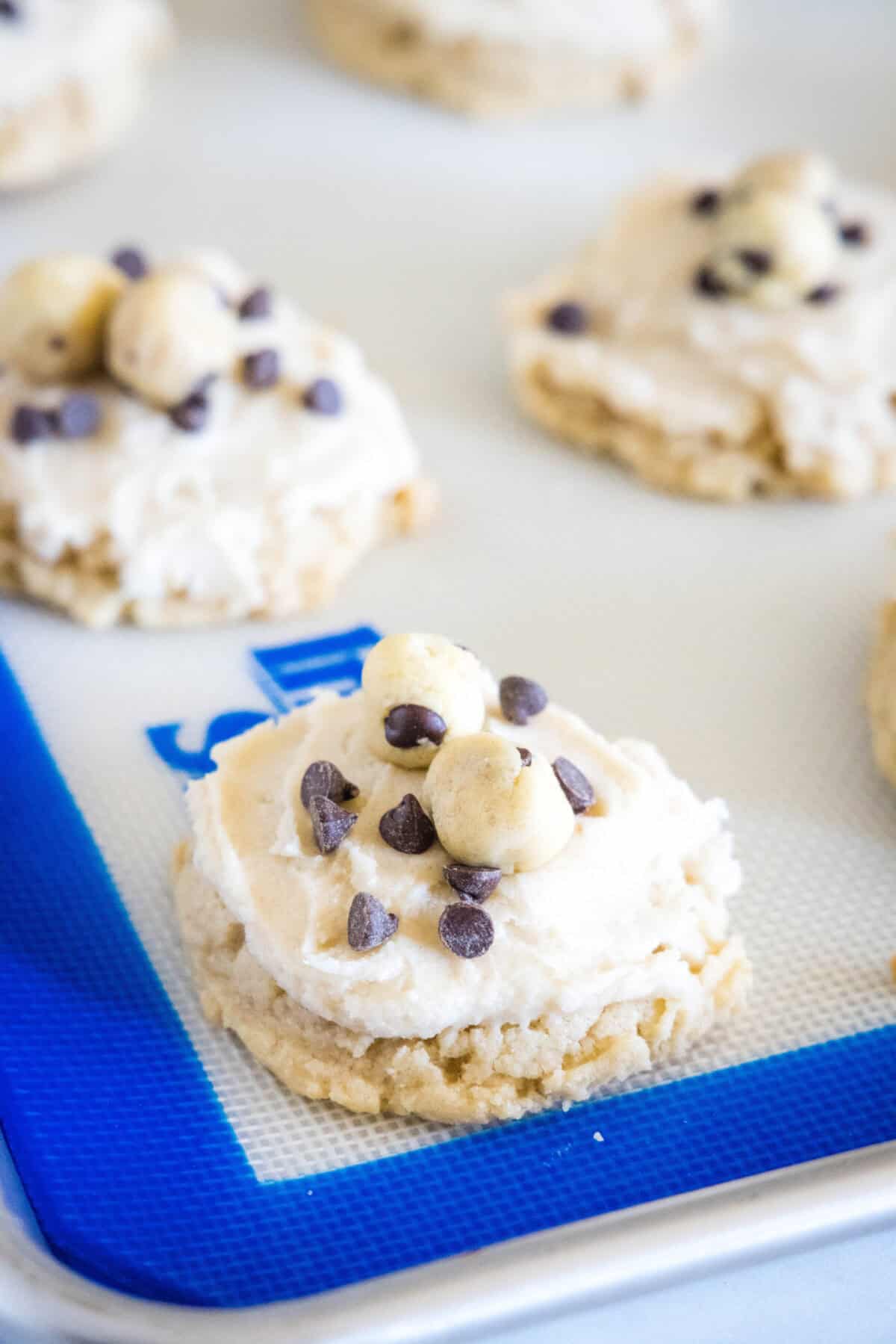 Cookie dough cookies frosted and topped with edible cookie dough and mini chocolate chips on a silpat-lined baking sheet.