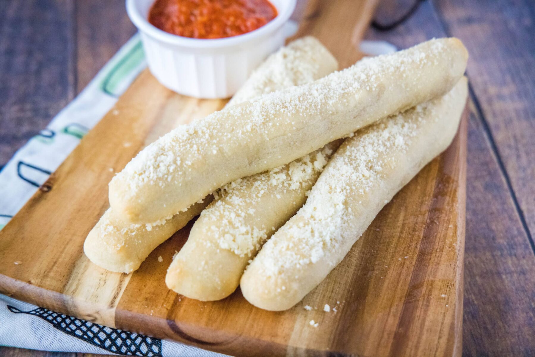 Four breadsticks in a pile on a cutting board in front of a ramekin of marinara
