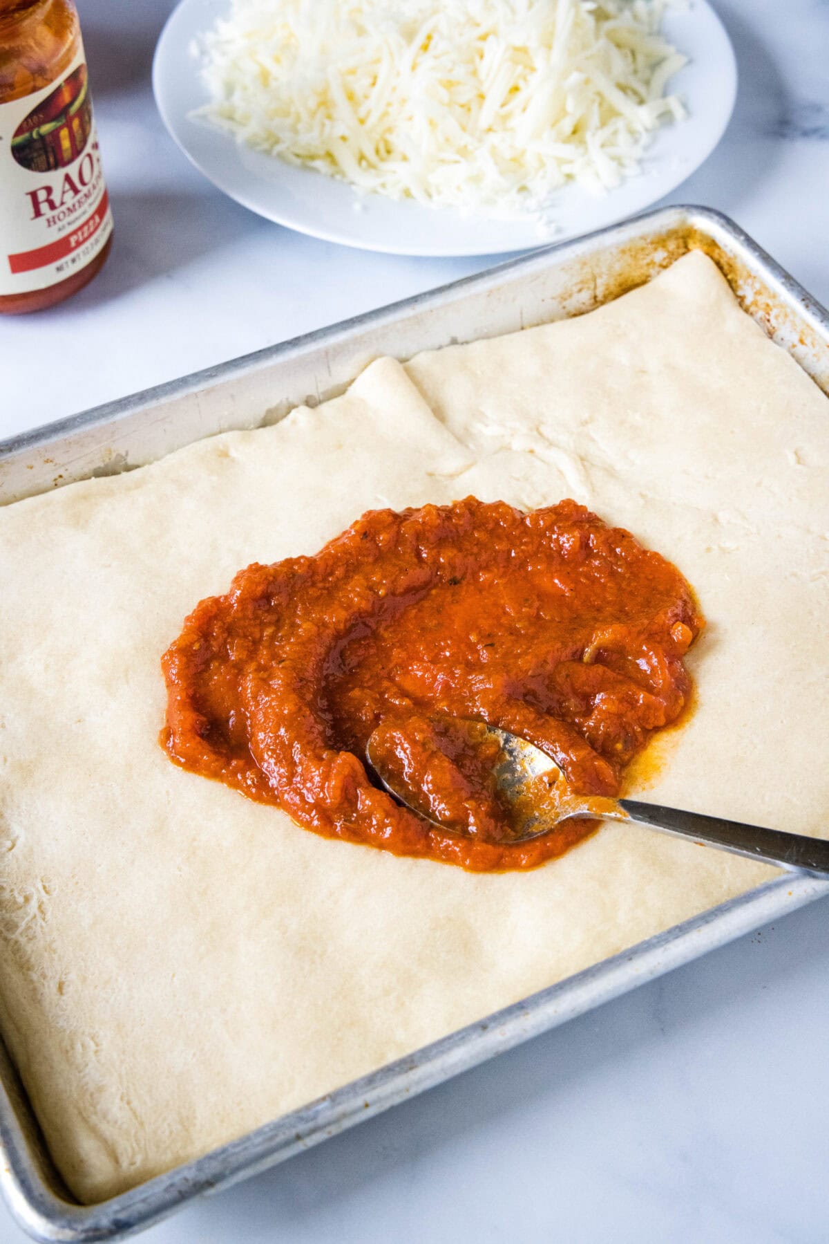 A spoon resting in a circle of partially spread pizza sauce in the middle of a crescent dough pizza crust.