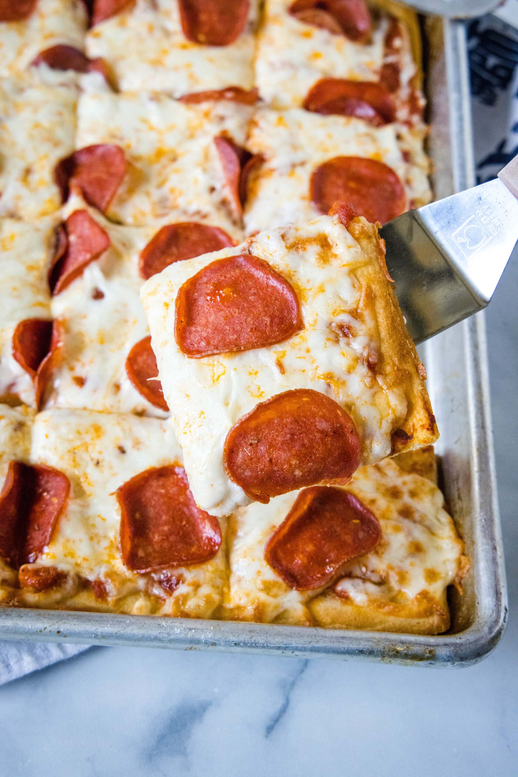 A spatula holding a pepperoni pizza slice over the rest of the crescent roll pizza on a baking sheet.