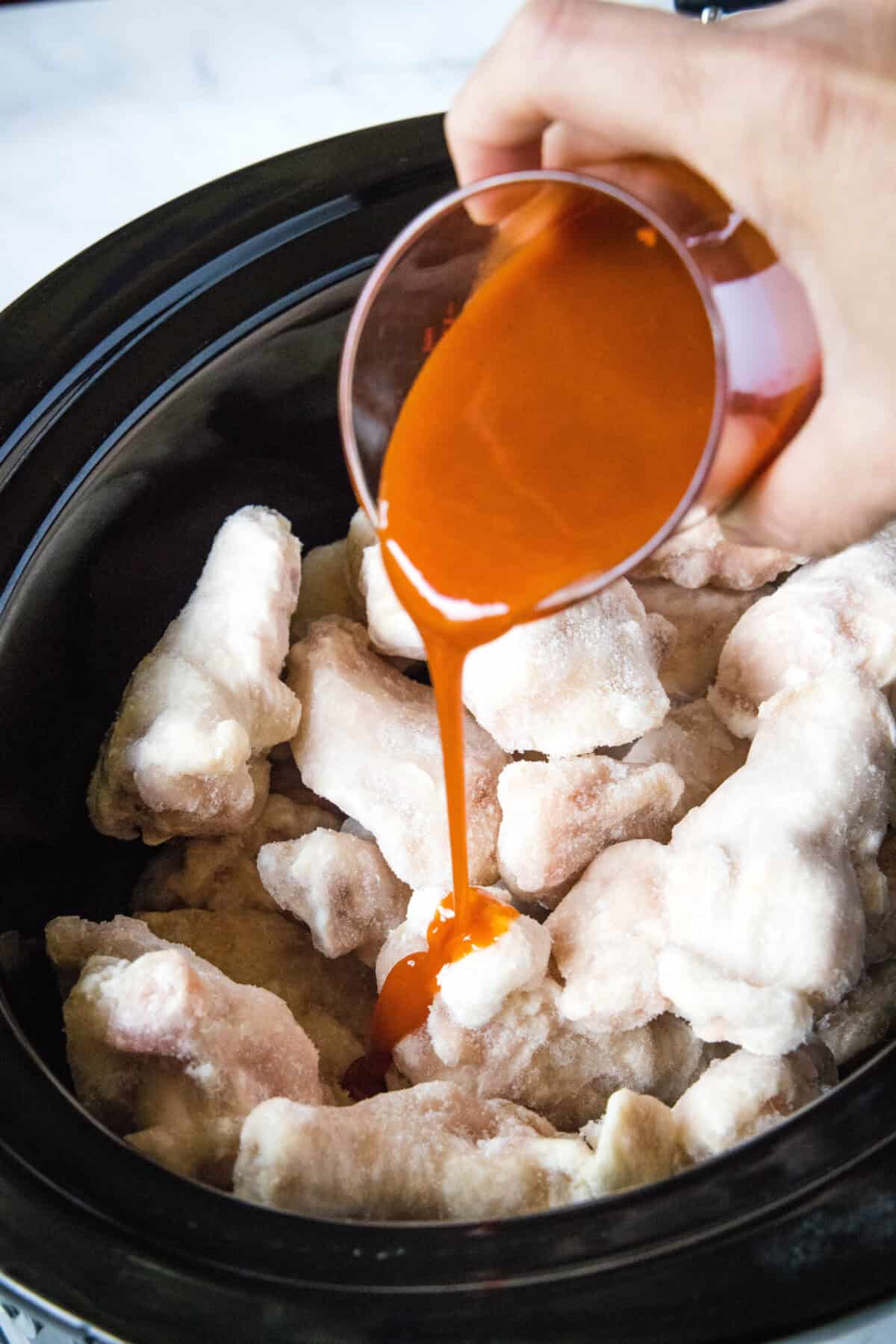 pouring buffalo sauce over chicken wings in a crockpot