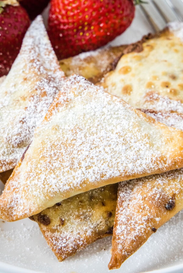 Close up of a pile of dessert wontons covered in powdered sugar next to strawberries and a fork