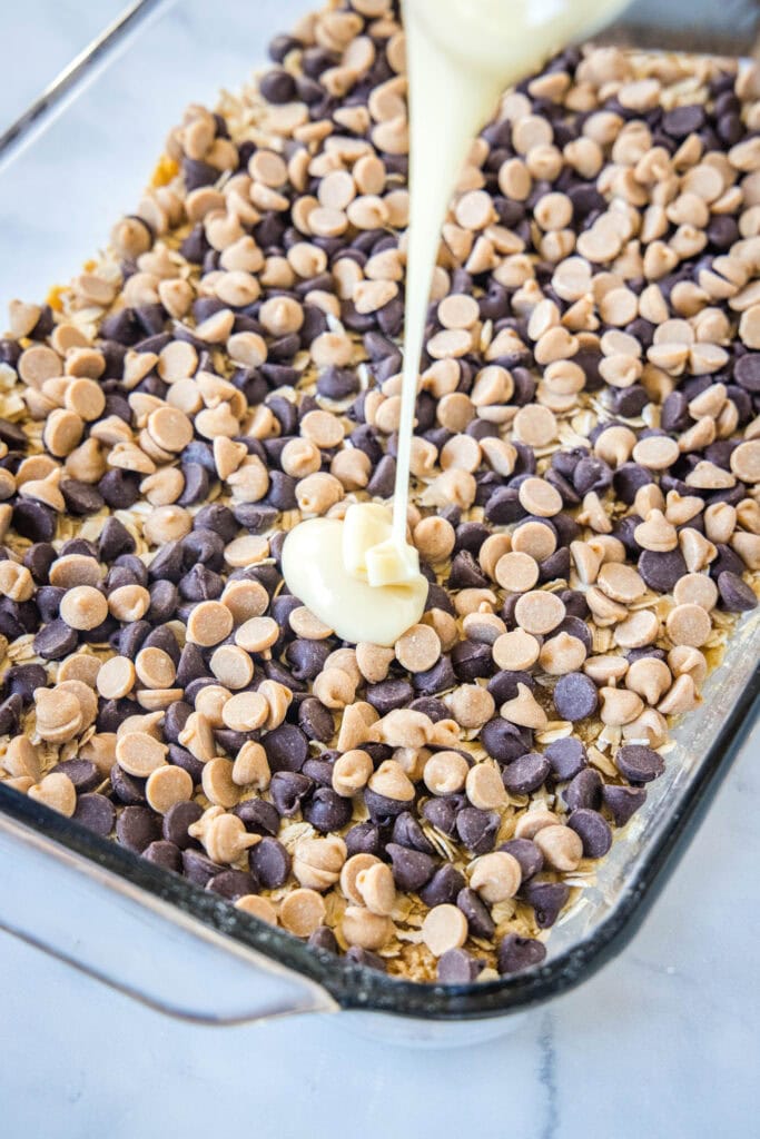 pouring sweetened condensed milk over peanut butter and chocolate chip layer of hello dolly bars