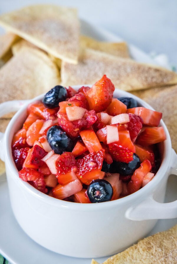 Fruit salsa in a white ceramic serving bowl with two handles, surrounded by cinnamon chips.