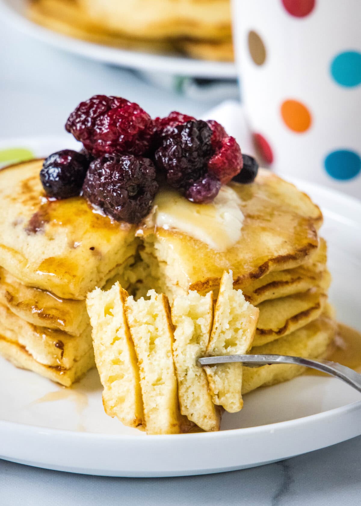 A forkful of pancakes resting in front of a stack of griddle cakes topped with mixed berries and syrup on a white plate.
