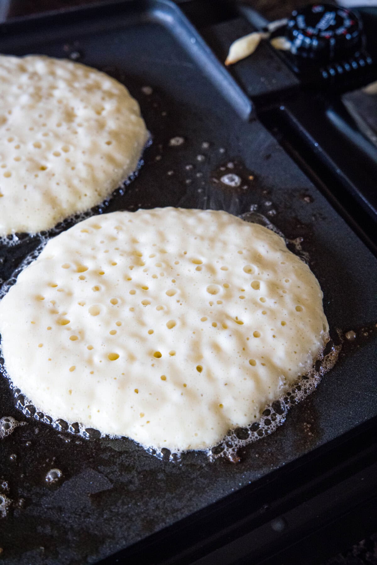 Griddle cake batter cooking on a skillet, with bubbles forming on the surface of the pancake.