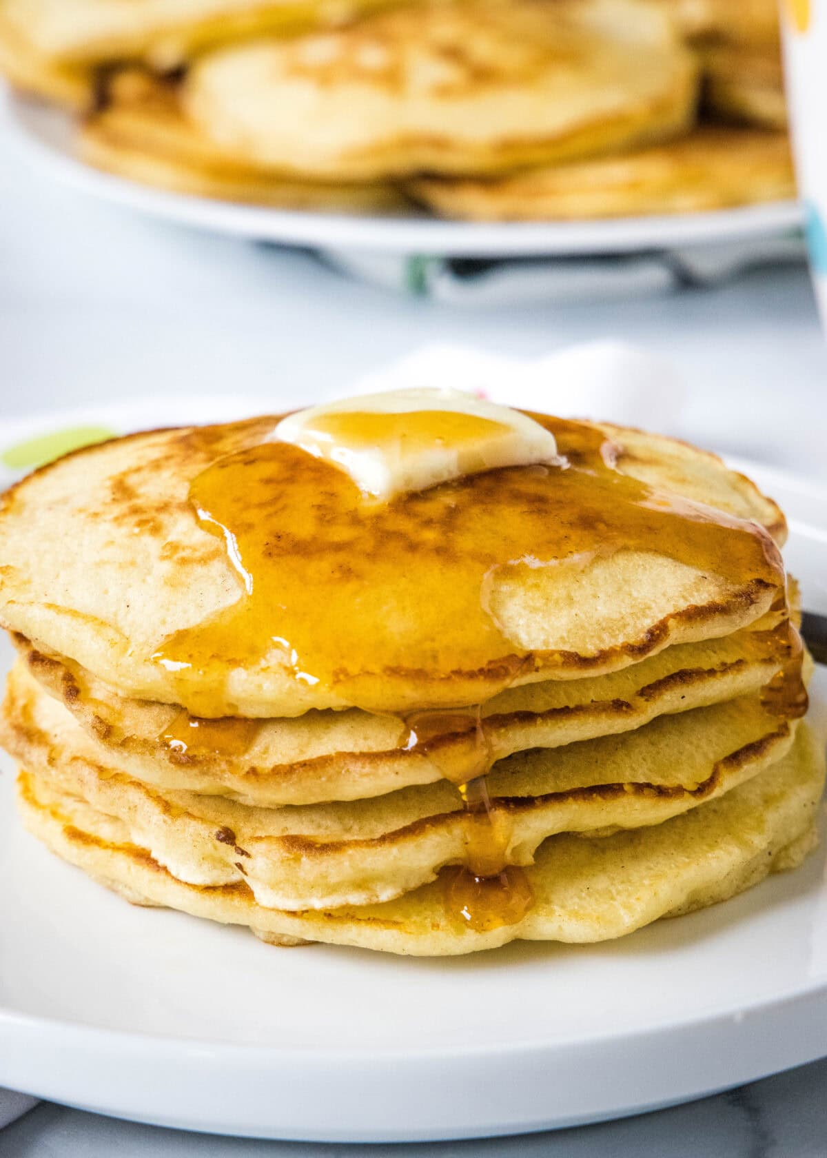 A stack of griddle cakes topped with a pat of butter and syrup on a white plate, with more pancakes on a platter in the background.