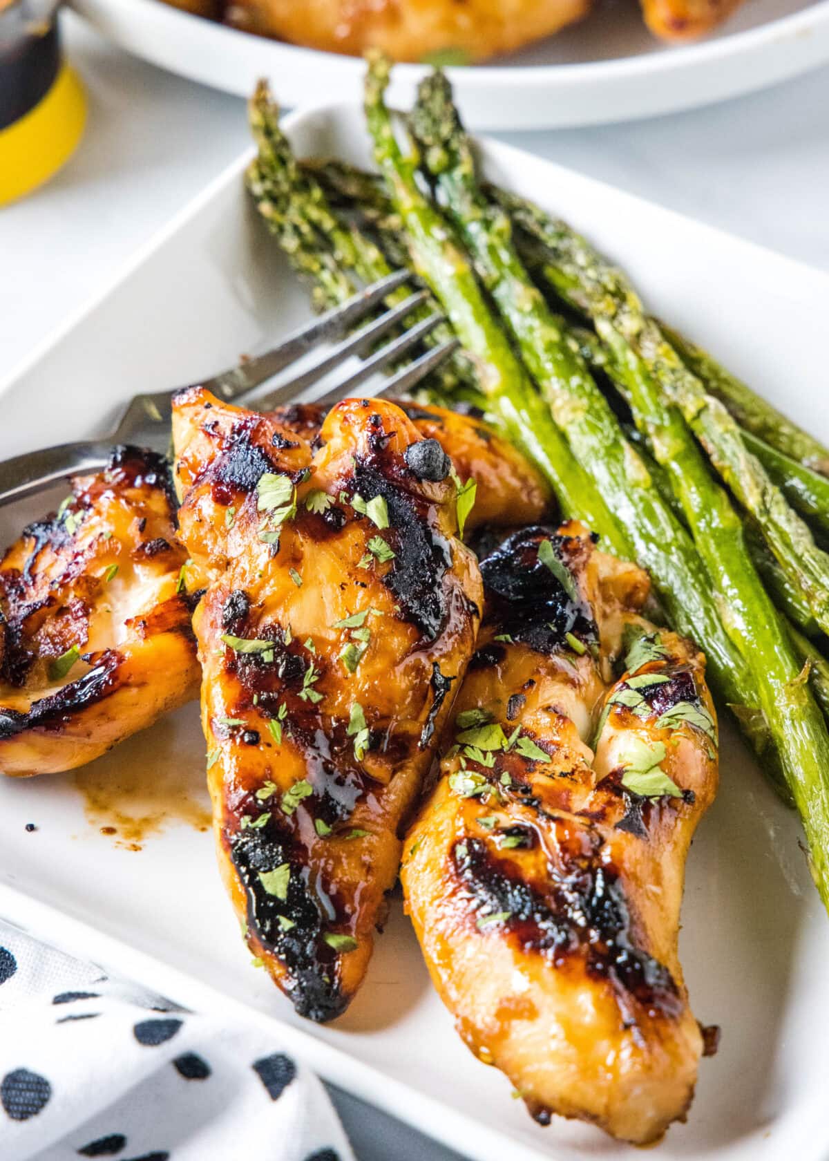 Grilled teriyaki chicken on a square plate next to a side of asparagus and a fork.