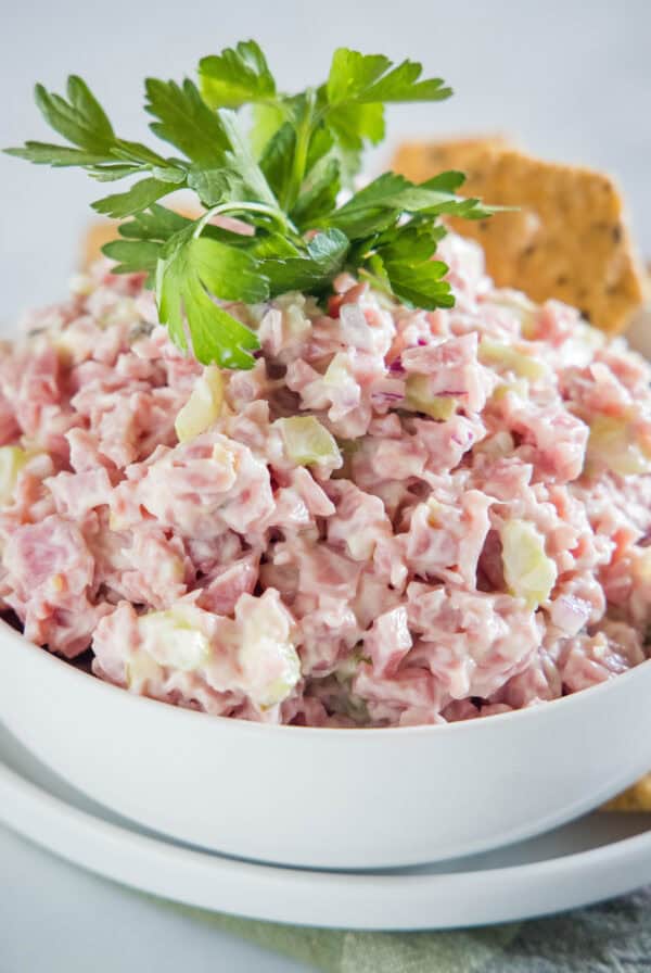 Ham salad in a white bowl garnished with a cracker and fresh parsley leaves.