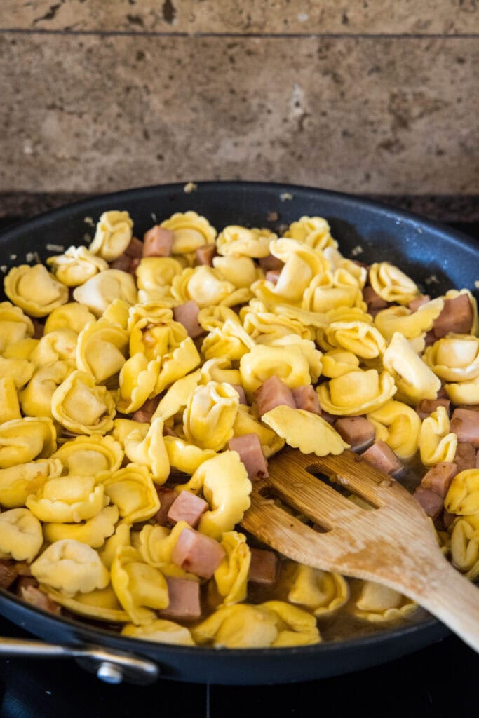 A wooden spatula stirring together cheese tortellini, diced ham, and onions in a skillet.