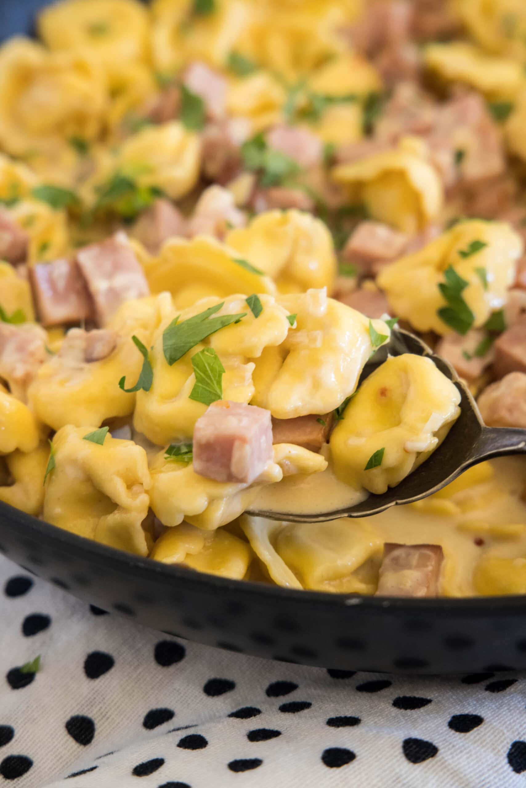 Close up of a serving spoon scooping ham tortellini from a large skillet.