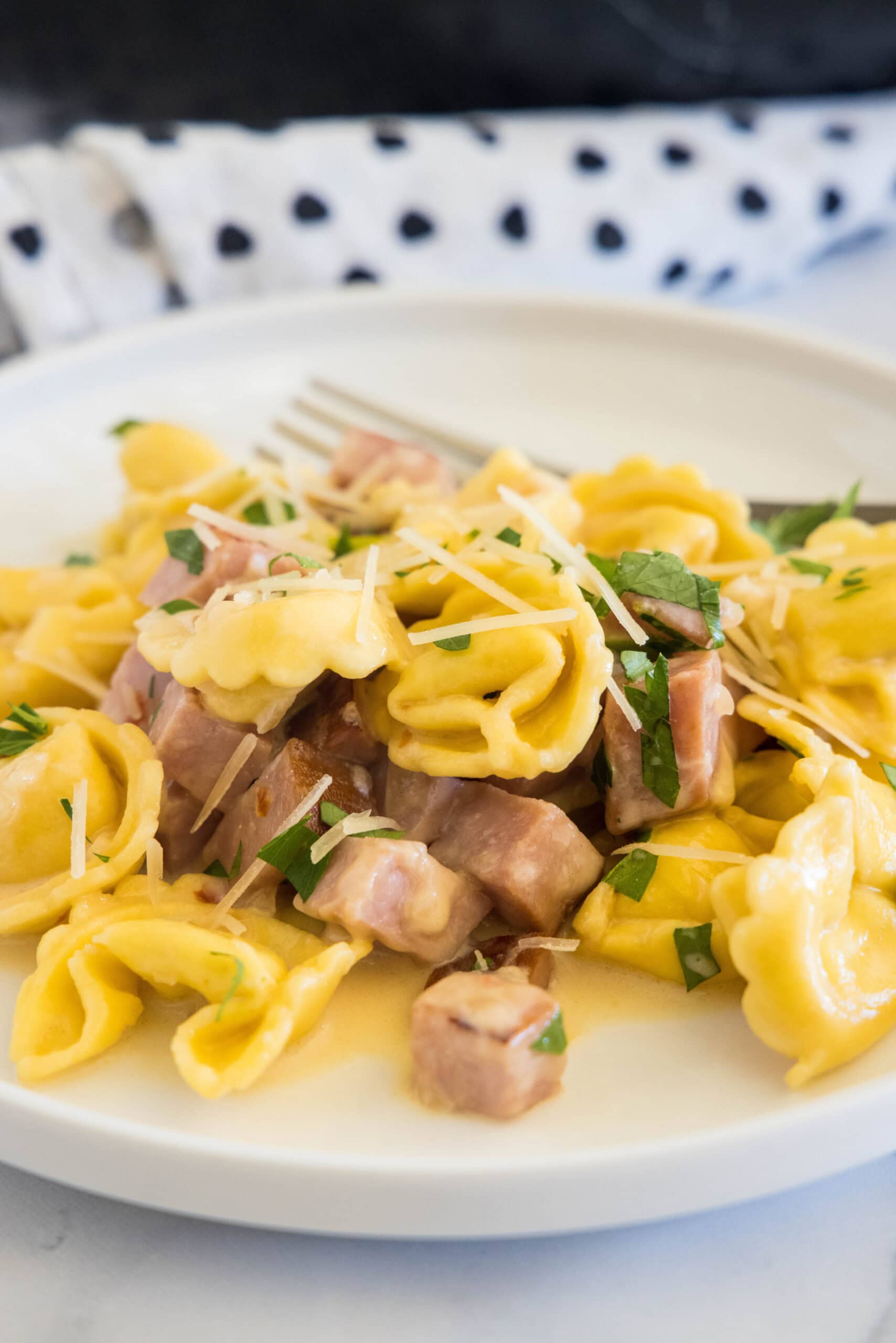 Ham tortellini garnished with chopped parsley and parmesan on a white plate, with a fork in the background.