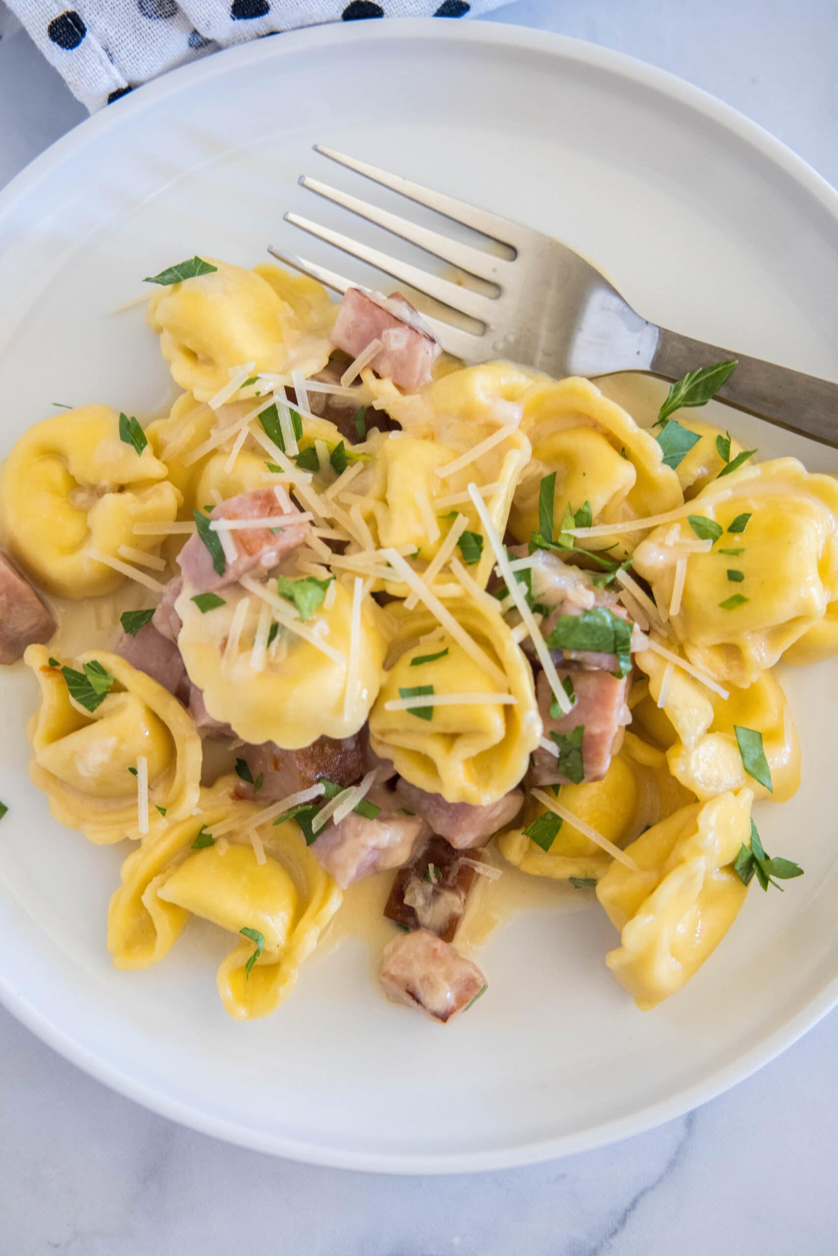 Overhead view of ham tortellini garnished with chopped parsley and parmesan on a white plate, next to a fork.