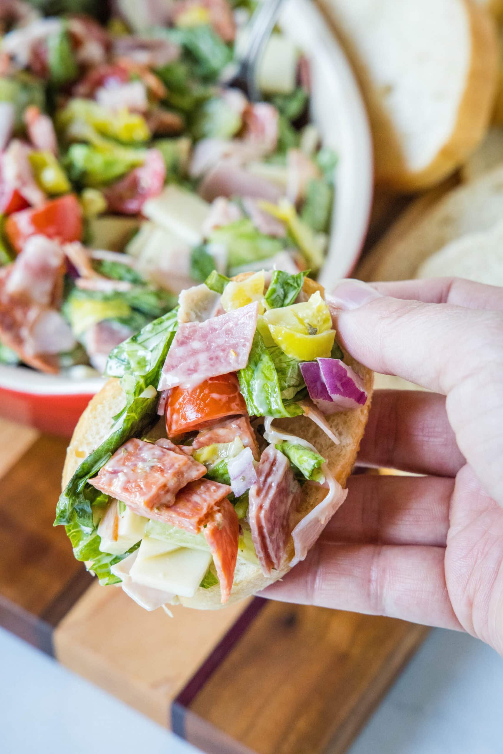 A hand holding up a bread slice topped with hoagie dip, with the bowl of dip and more bread slices in the background.