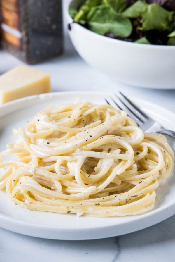 A serving of pasta alfredo on a white plate next to a fork, with a bowl of salad in the background.