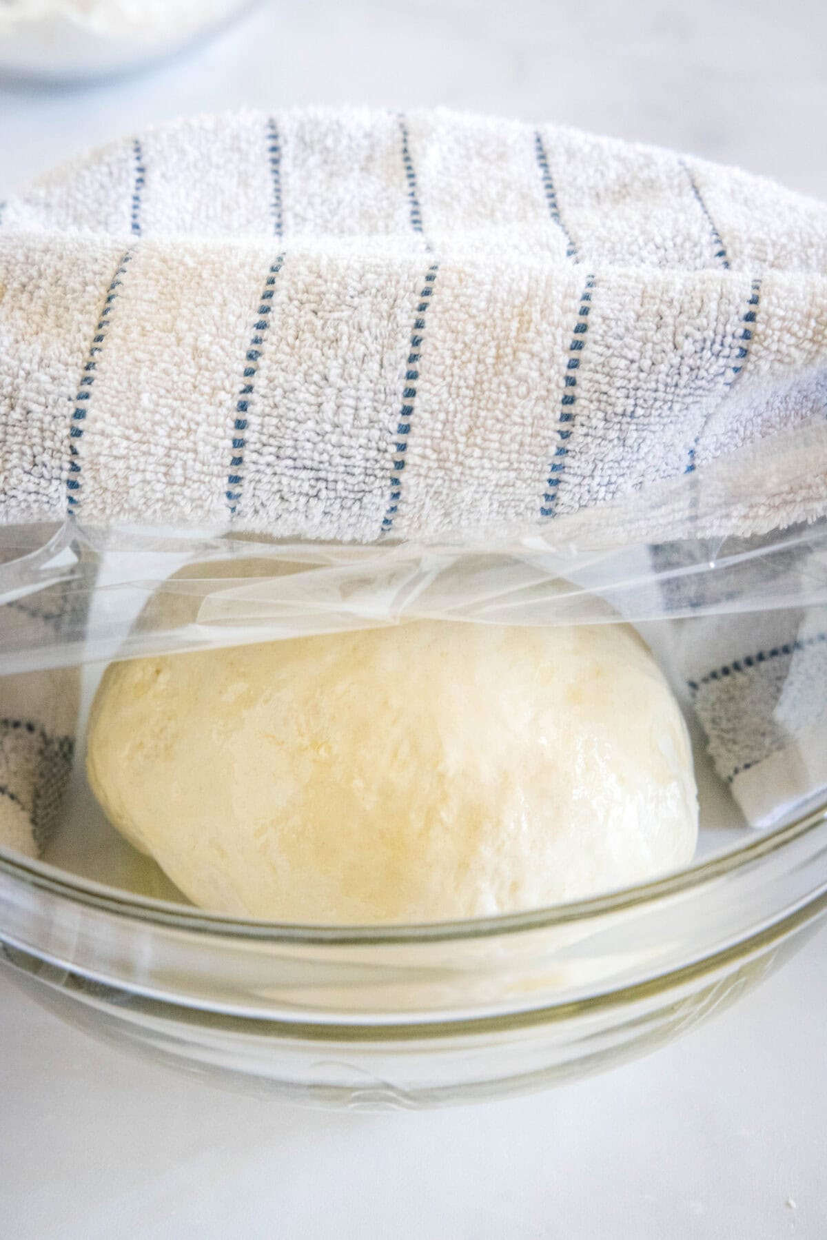 A ball of pita bread dough in a glass bowl covered with plastic wrap and a white and blue striped dishcloth.