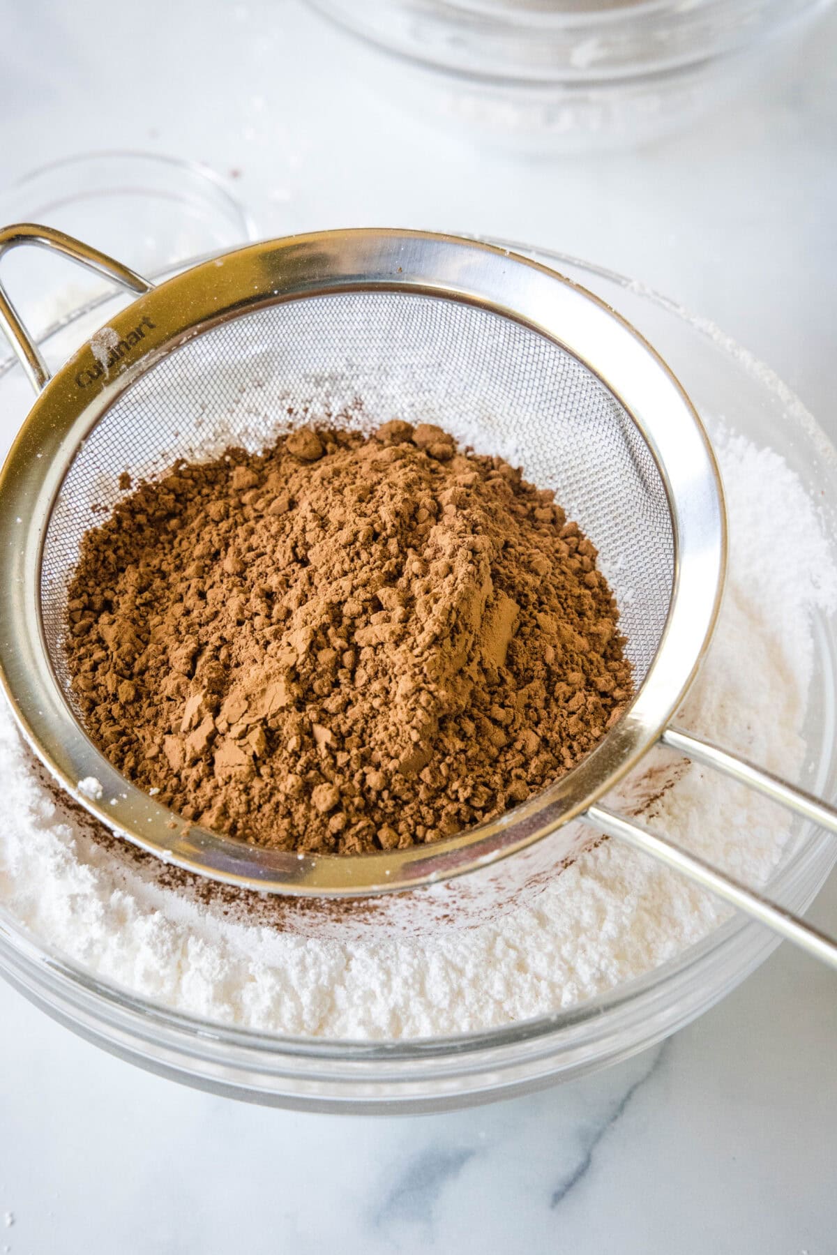 Cocoa powder in a sifter held over sugar in a glass bowl.