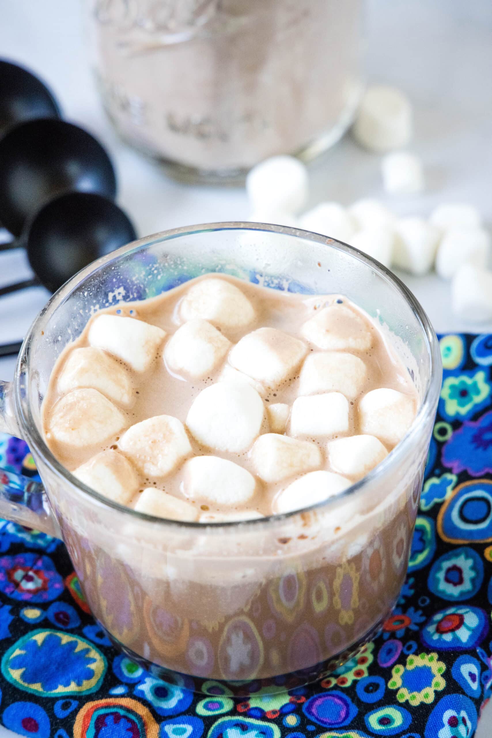 Hot chocolate in a clear mug topped with mini marshmallows, with more mini marshmallows and a jar of hot chocolate mix in the background.