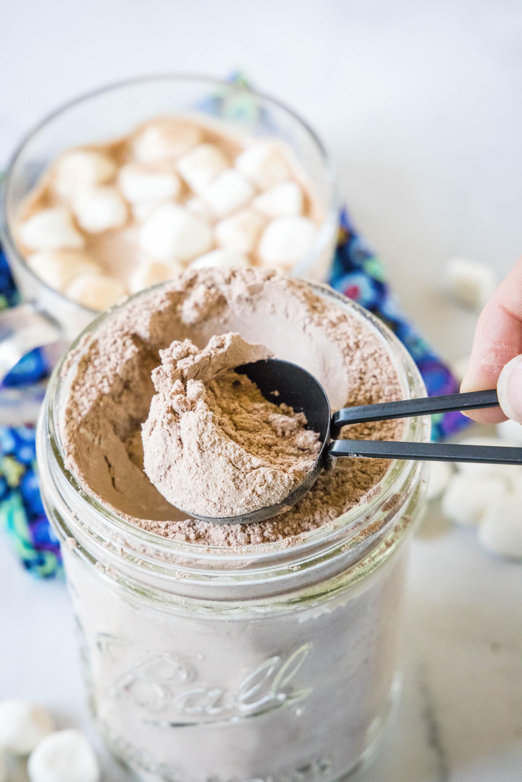 A tablespoon scooping hot chocolate mix from a glass jar, with a muf og hot cocoa in the background.