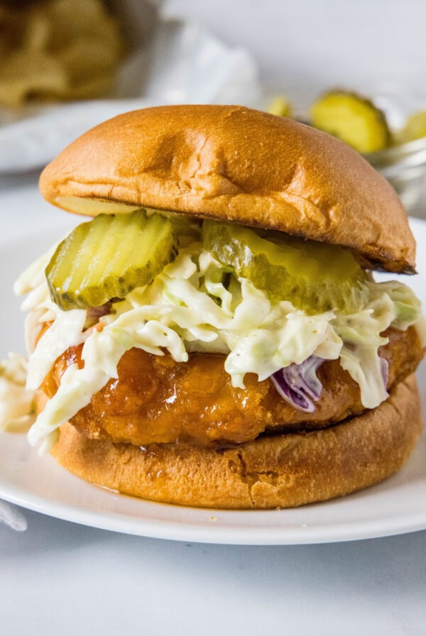 A hot honey chicken sandwich topped with coleslaw and pickles on a white plate.