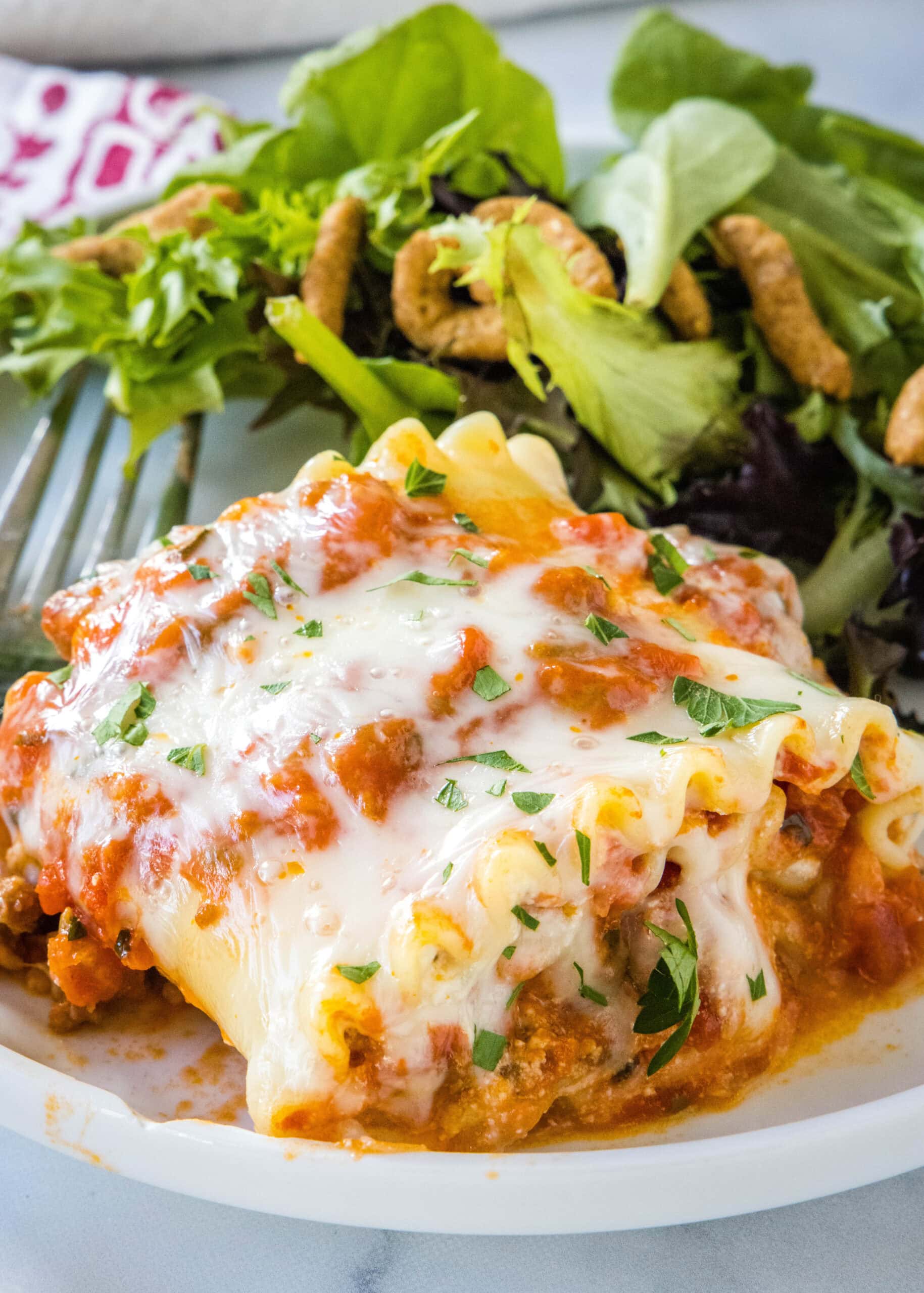 Lasagna roll ups are a fun spin on classic lasagna. This recipe is packed with savory meat sauce and melty cheese, and it's easy to customize! A lasagna roll up served on a plate with green salad on the side, next to a fork.
