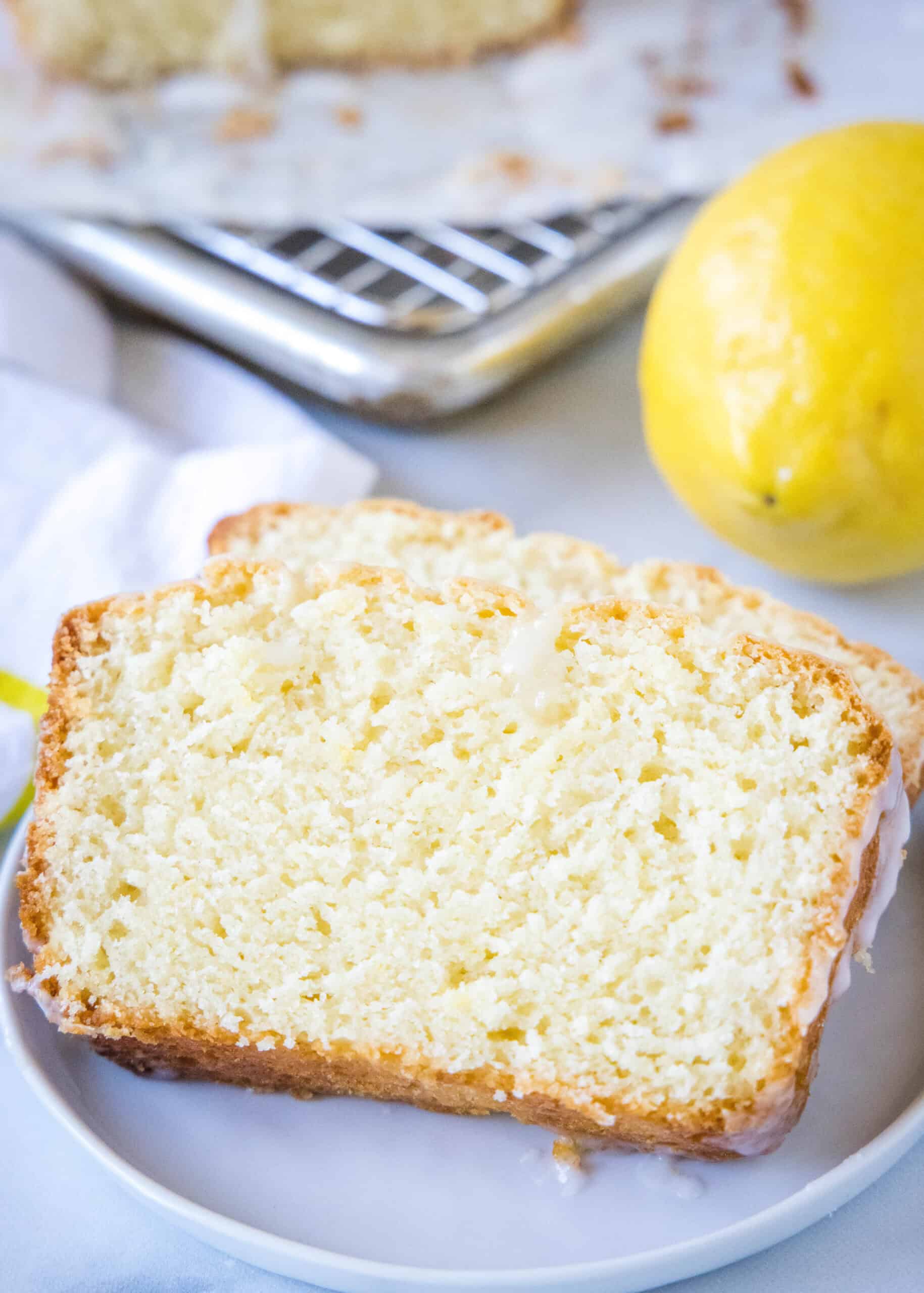Two lemon loaf slices on a white plate with a fresh lemon in the background.