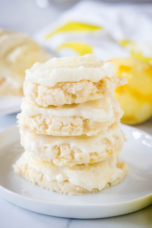 Four frosted lemon sugar cookies stacked on a plate with a zested lemon in the background.