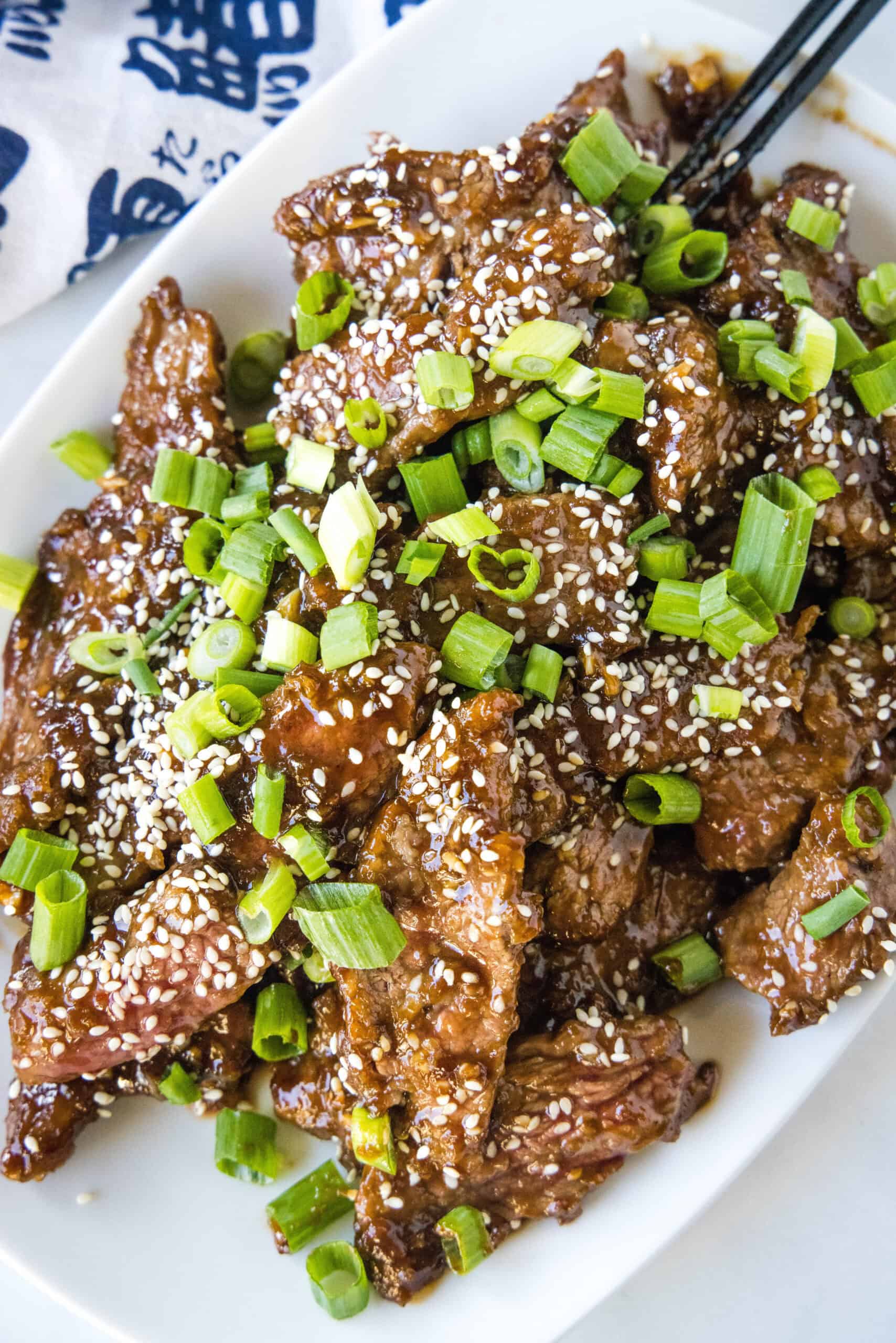 Overhead view of Mongolian beef topped with sesame seeds and green onions on a white platter.