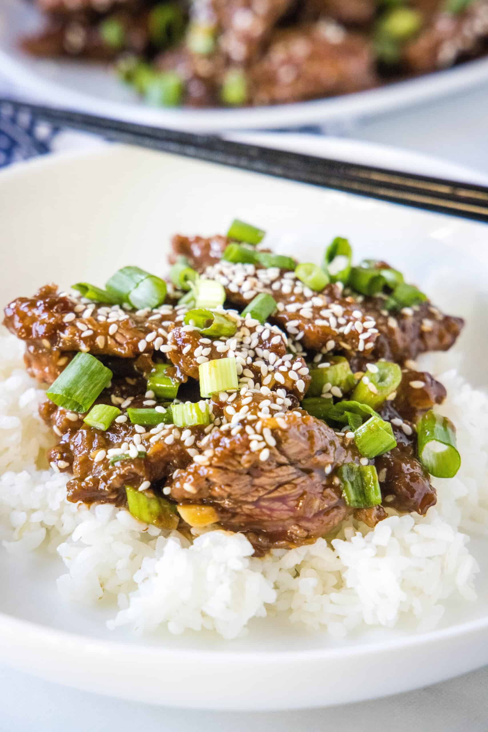 Mongolian beef served over rice, topped with sesame seeds and green onions in a white bowl with chopsticks resting in the background.
