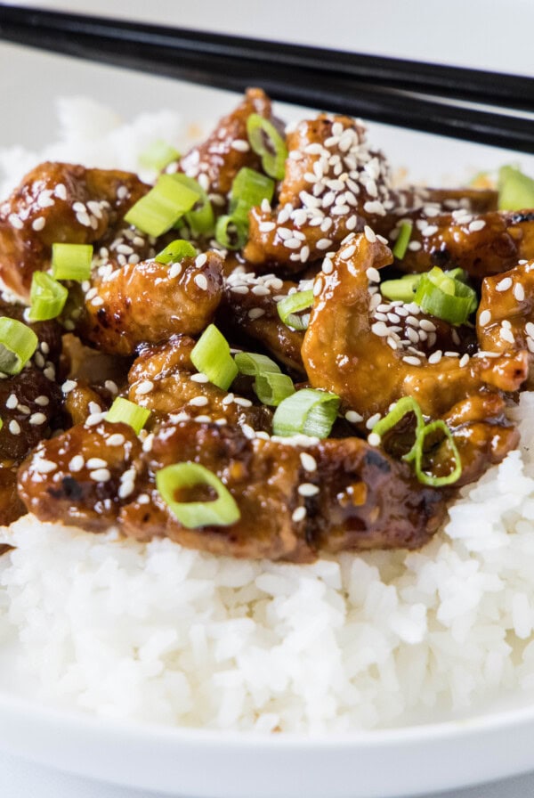 A plate with Mongolian pork on top of rice, topped with sesame seeds and green onions, with a pair of chopsticks.