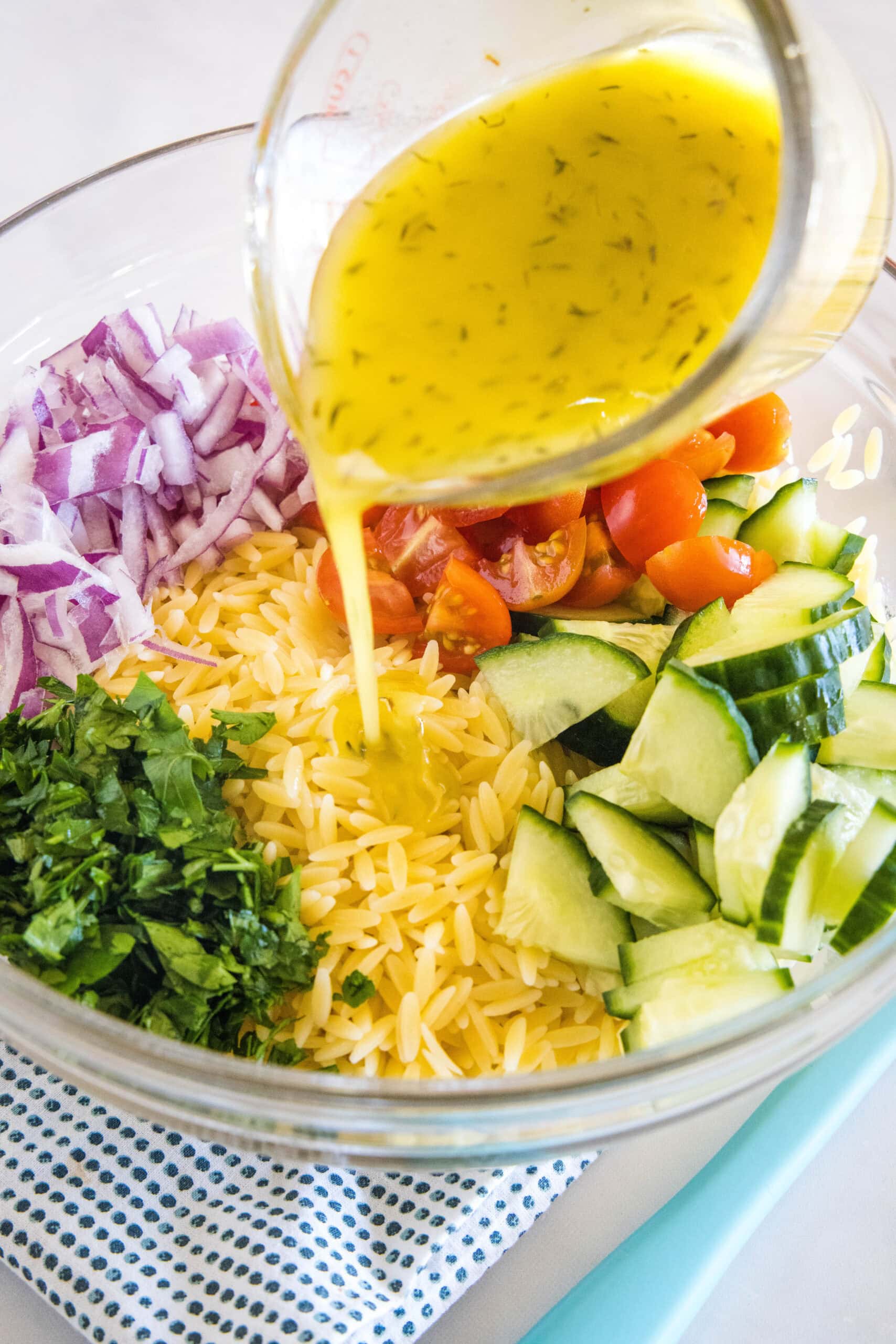 Lemon vinaigrette being poured from a measuring cup over orzo pasta salad ingredients in a large glass bowl.