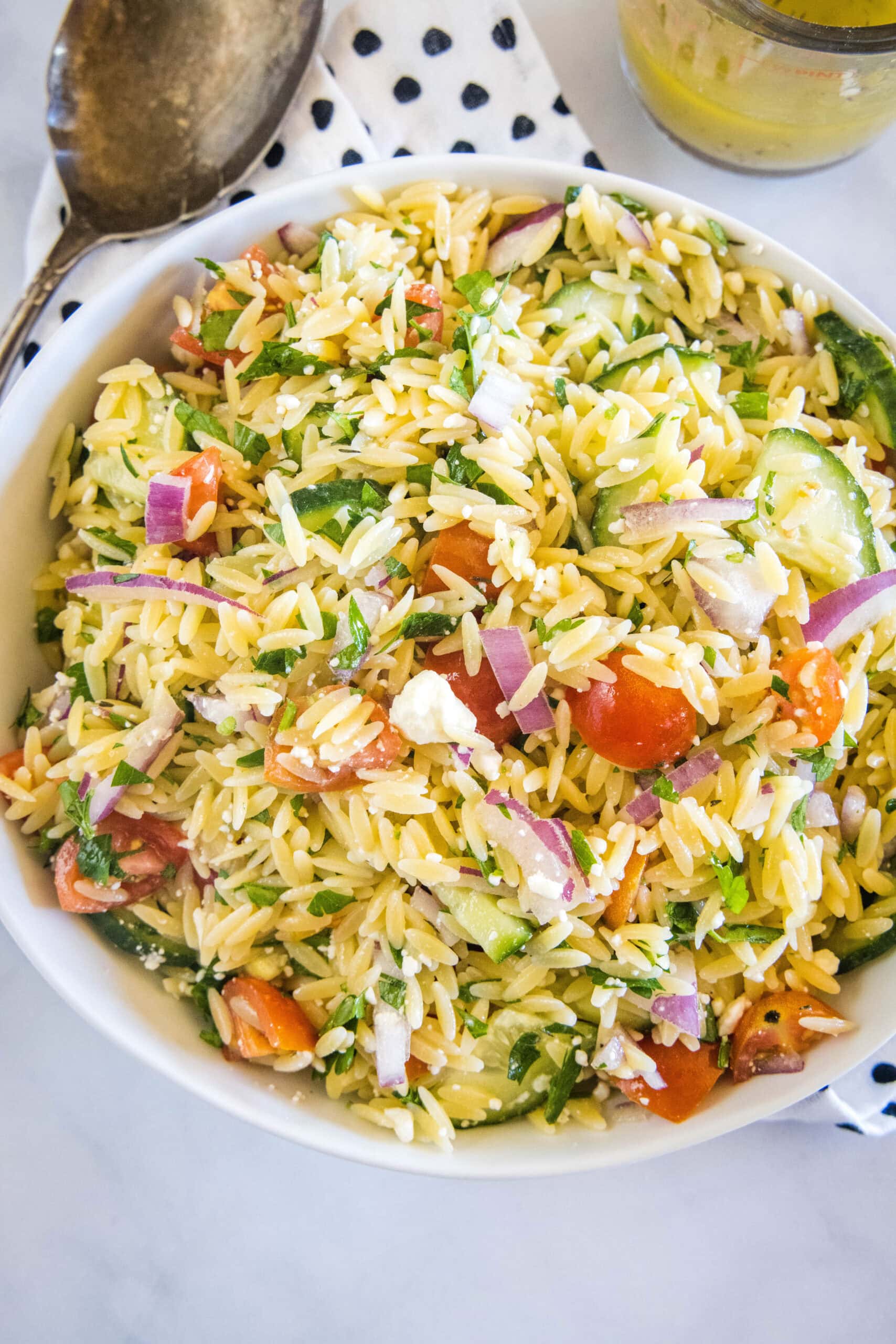 Overhead view of orzo pasta salad in a large bowl next to a serving spoon and a jar of dressing.