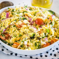 Orzo pasta salad in a large bowl resting on top of a black and white polka dot dishcloth, with a jar of dressing in the background.