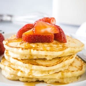 A stack of homemade pancakes topped with fresh strawberries and syrup on a white plate.