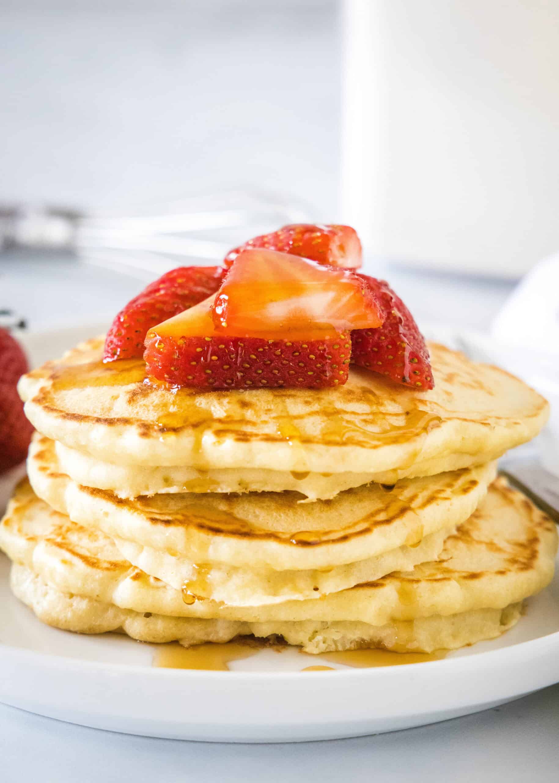 A stack of homemade pancakes topped with fresh strawberries and syrup on a white plate.