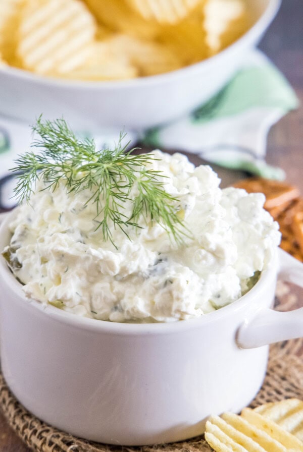 Pickle dip topped with fresh dill sprigs in a white serving dish, with a bowl of chips in the background.
