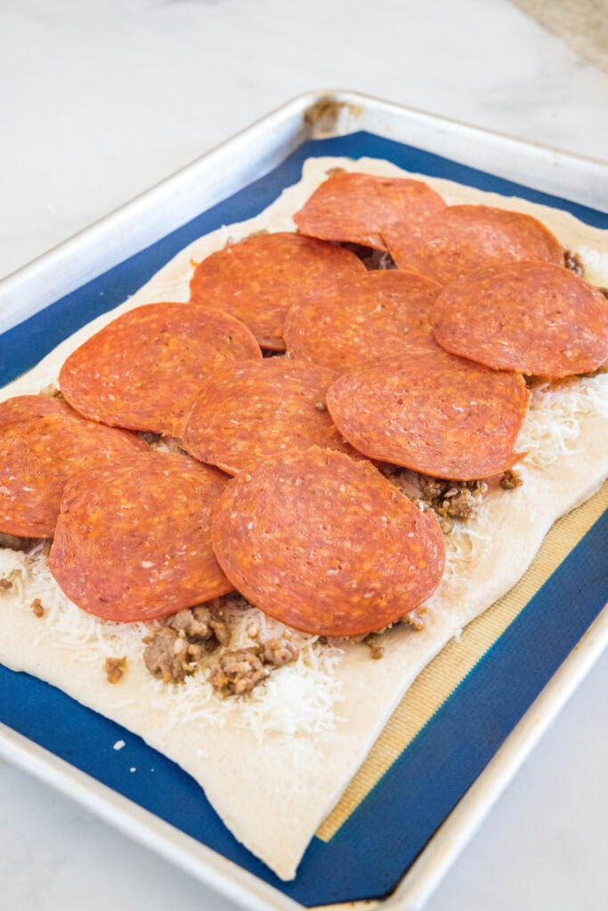 Pizza dough topped with shredded cheese, ground meat, and pepperoni slices on a baking sheet.