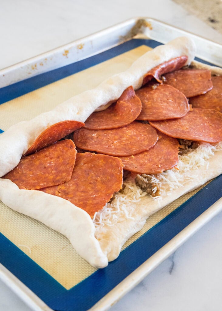 Pizza dough filled with toppings and partially rolled into a log on a baking sheet.