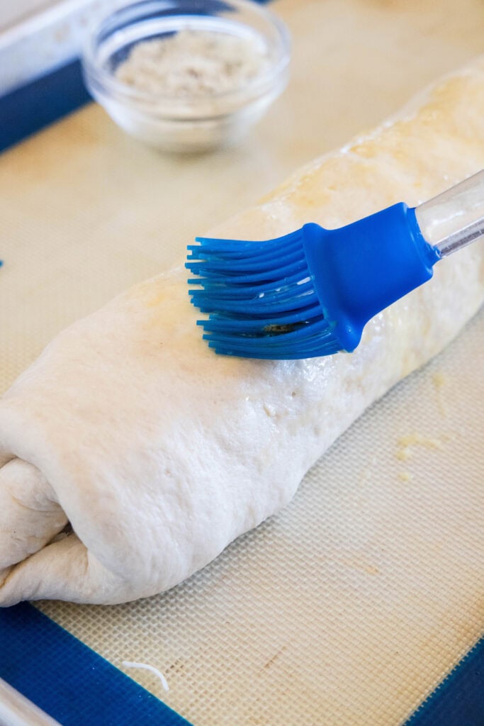 A blue basting brush brushing an egg wash over the top of rolled up pizza dough on a baking sheet.