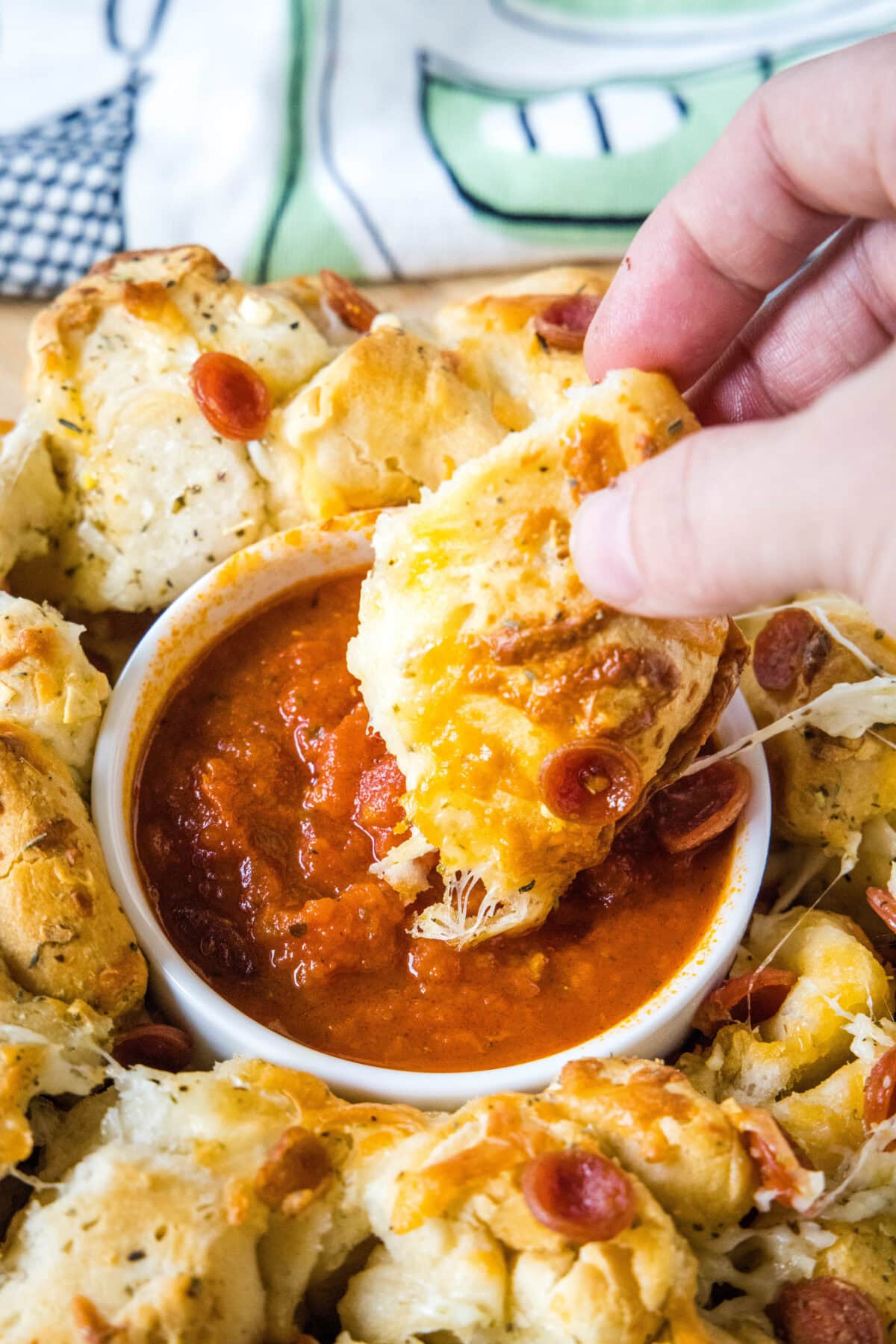 A hand dipping a piece of pizza monkey bread into a bowl of pizza sauce surrounded by the rest of the monkey bread.